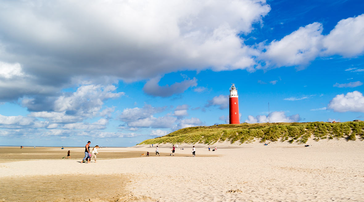 Large stretch of sand under big blue sky with a bright red lighthouse on top of a green dune